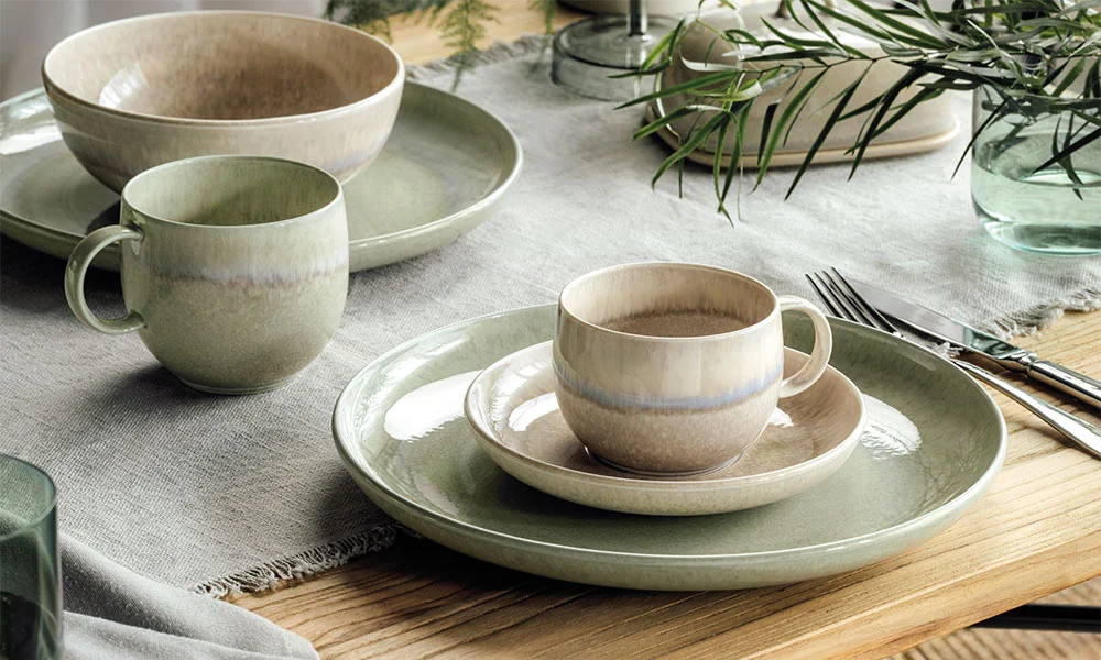Keramikgeschirr-Set der Serie Perlemor von Villeroy &amp; Boch in sanften Grüntönen auf hölzernem Tisch mit Leinentuch, dekoriert mit Pflanzen.