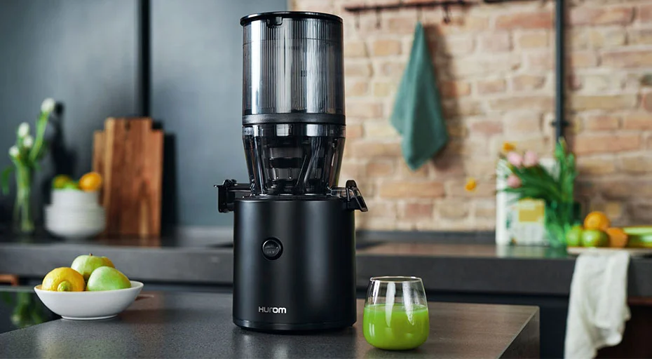 Schwarzer Hurom-Entsafter auf Küchenarbeitsplatte mit Glas grünen Saft und Obst im Hintergrund.