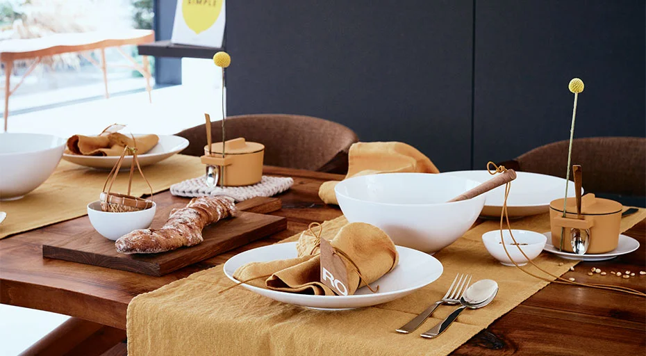 Eleganter Esstisch mit Brot, Tellern, Schalen  der Serie TAC Gropius weiss von Rosenthal und gelben Servietten, dekoriert mit minimalistischen Blumenarrangements.