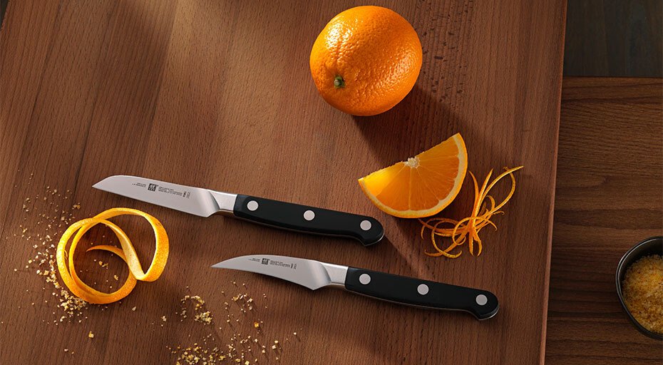 Zwei Schälmesser mit schwarzen Griffen, eine ganze Orange und geschnittene Schale auf einem Holzbrett.