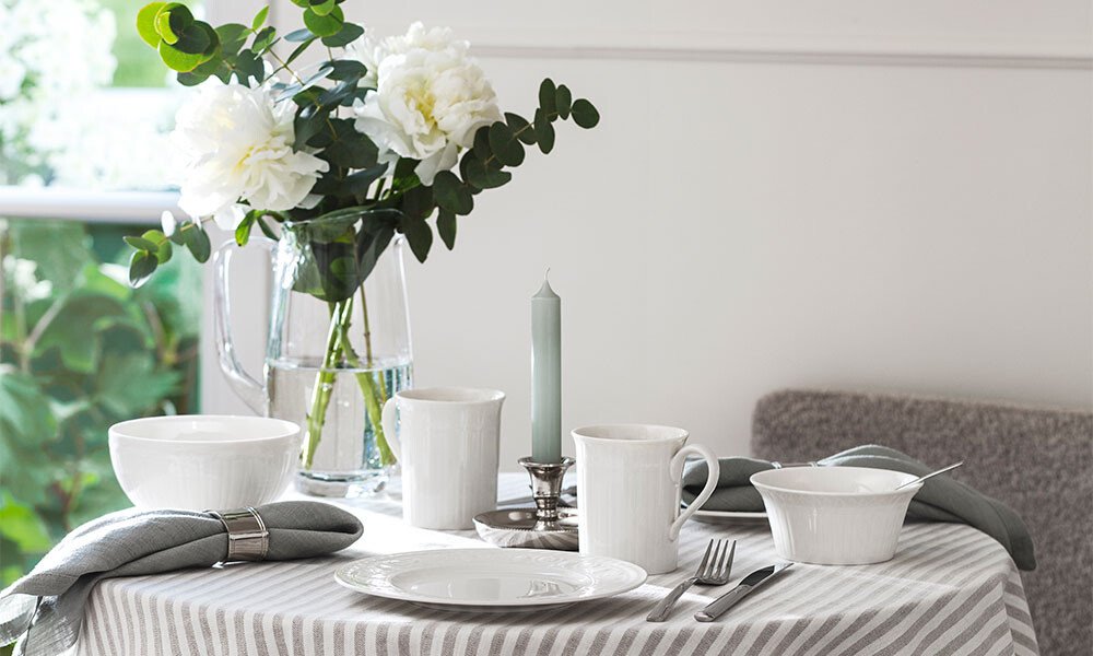 Gedeckter Tisch mit gestreifter Tischdecke, weißem Geschirr der Serie Cellini von Villeroy &amp; Boch, Kerze und Blumenstrauß in Vase.