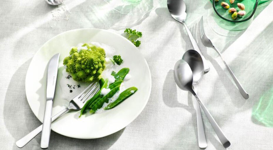 Weißer Teller mit Romanesco, Erbsen, Kräutern und Besteck der Serie Atlantic von Robbe & Berking auf heller Tischdecke, Pistazien in grünem Glas.