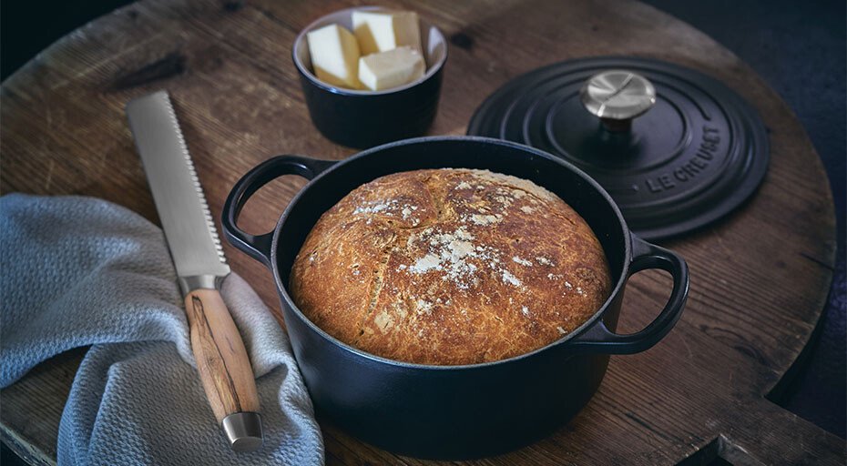 Brotlaib in einem gusseisernen Bräter von Le Creuset neben einem Brotmesser, Butter und einem Tuch auf einem Holztisch.