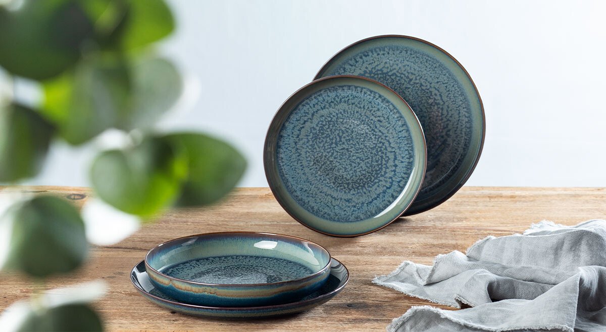 Geschirr-Set, bestehend aus zwei blauen Tellern auf Holztisch, links unscharfe Pflanze, rechts graues Tuch. Minimalistische Ästhetik.