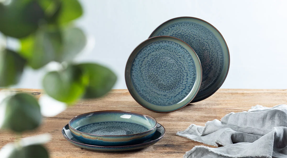 Geschirr-Set, bestehend aus zwei blauen Tellern auf Holztisch, links unscharfe Pflanze, rechts graues Tuch. Minimalistische Ästhetik.