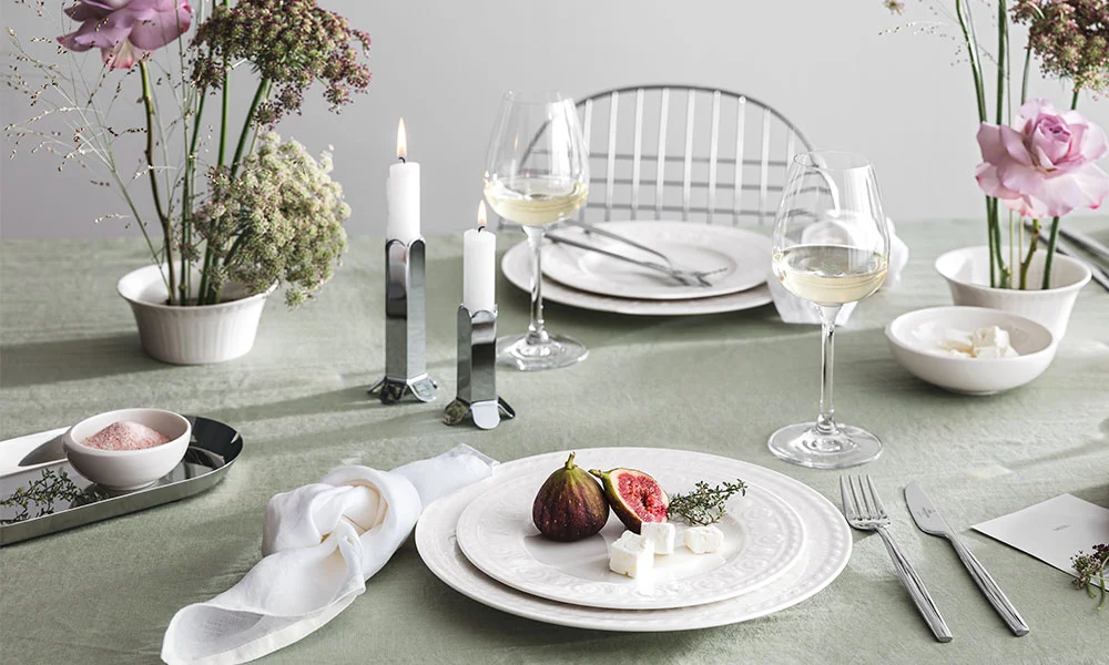 Eleganter gedeckter Tisch mit Feigen, Käse, Weingläsern und Blumenarrangement auf grünem Tischtuch.