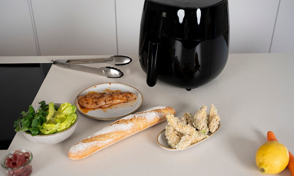 Airfryer auf einer Küchenarbeitsplatte mit Baguette, Salat, Hähnchenbrust, Gemüse und einer Zange.