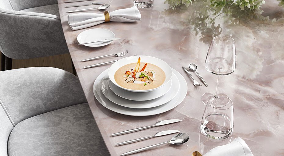 Elegante Tischdekoration mit Suppenteller, Besteck, Gläsern und Blumenarrangement auf einem polierten Marmortisch.