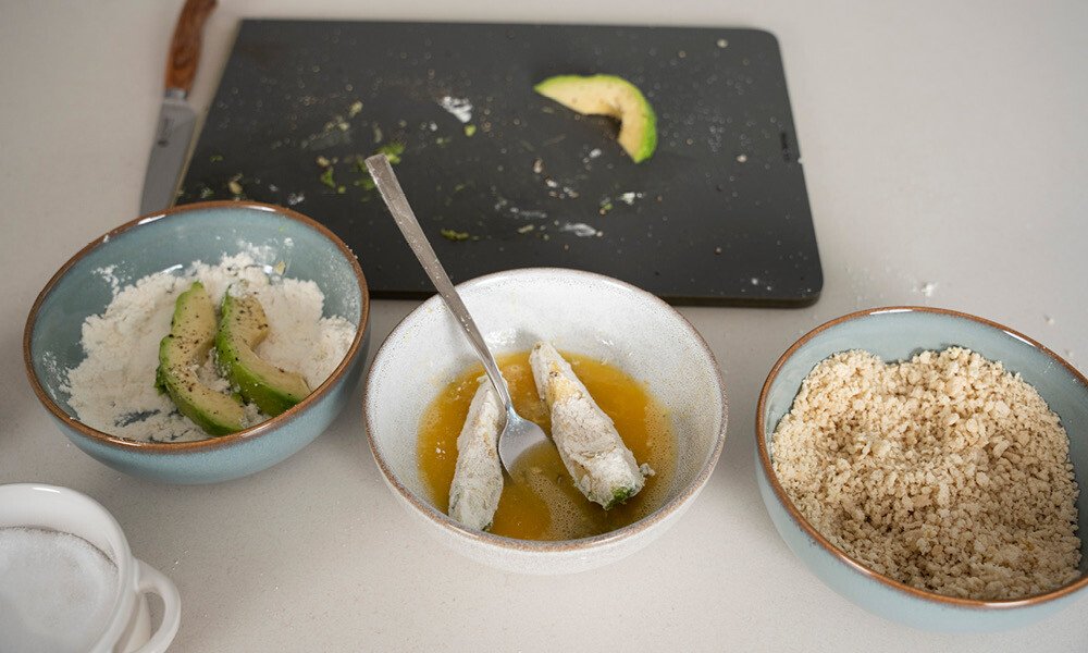 Drei Schüsseln mit Zutaten: Mehl und Avocadoscheiben, Ei-Mischung, Paniermehl; Schneidebrett mit Avocadoresten im Hintergrund.
