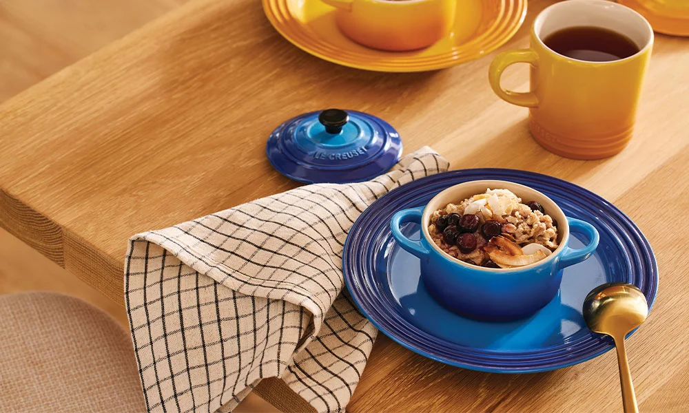 Kleiner blauer Topf mit Müsli, Nüssen und Beeren auf einem Teller, daneben Tasse Tee und Stoffserviette.
