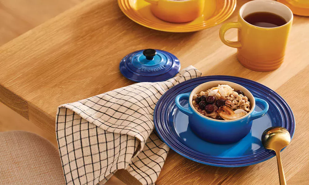 Kleiner blauer Topf mit Müsli, Nüssen und Beeren auf einem Teller, daneben Tasse Tee und Stoffserviette.