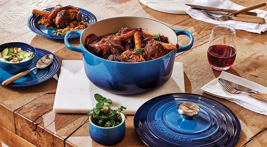 Blauer Bräter mit geschmortem Lamm, serviert auf Holztisch, begleitet von Salat, Rotwein und Kräutern in Schale.