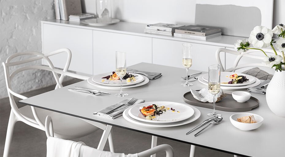 Eleganter, modern gedeckter Tisch mit Tellern der Serie NewMoon von Villeroy &amp; Boch, Besteck, Gläsern und Blumendekoration vor einem weißen Sideboard.