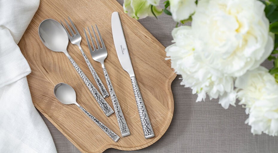 Besteckset der Serie Blacksmith von Villeroy & Boch mit texturierten Griffen auf Holzbrett, neben weißen Blumen arrangiert.