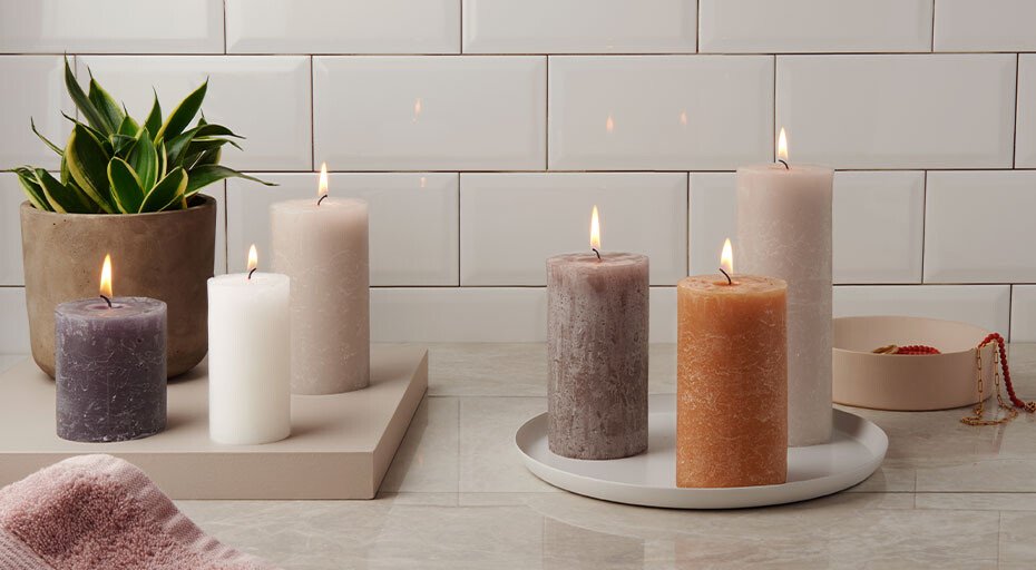 Verschiedene brennende Kerzen von Bolsius in Erdtönen auf einem Tablett vor einer Kachelwand, neben einer Pflanze und Schmuck.