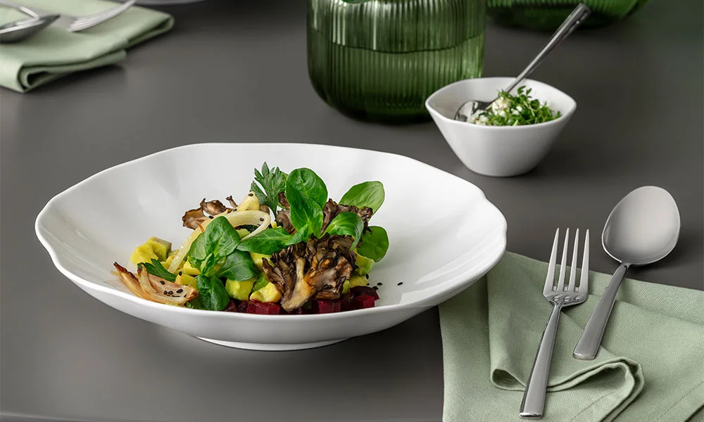 Elegantes Gericht mit Pilzen, Blattgrün und Gemüse auf weißem Teller der Serie Pura von Villeroy &amp; Boch, gedeckter Tisch im Hintergrund.