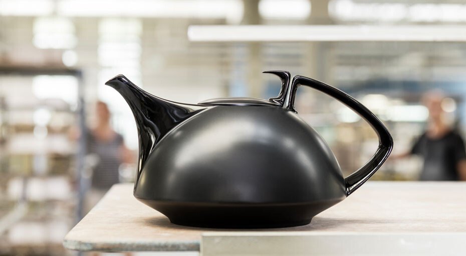 Moderne, schwarze Teekanne der Serie TAC Gropius black von Rosenthal mit dynamischem Design auf Tisch. Hintergrund unscharf mit Personen und Regalen.