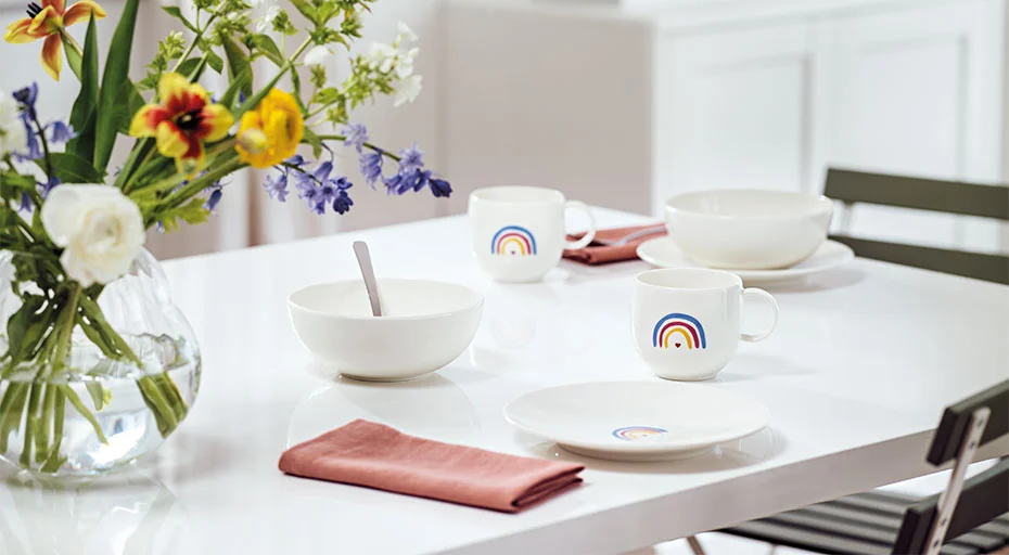 Ein gedeckter Tisch mit Villeroy &amp; Boch With Love Geschirr, bestückt mit Regenbogenmotiv. Vase mit Blumenarrangement daneben.