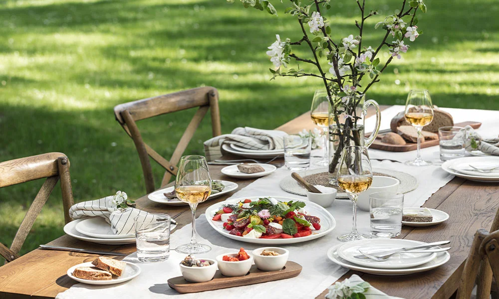 Eleganter Gartentisch mit festlichem Gedeck, frischem Salat, Brot, Weingläsern und Blumenarrangement unter blühendem Baum.