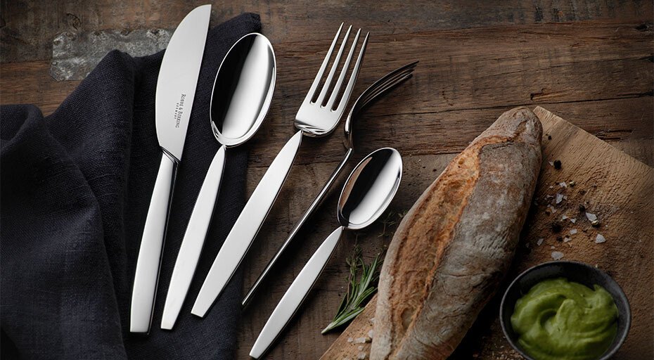 Besteck-Set der Kollektion 12&quot; von Robbe &amp; Berking neben Baguette und Schale mit grünem Dip auf Holzuntergrund.
