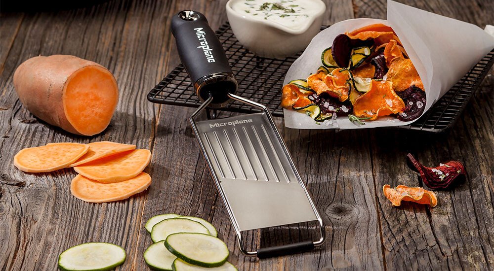Gemüsehobel der Gourmet-Serie von Microplane auf Holzbrett mit geschnittenen Süßkartoffeln und Zucchini. Chips und Dip im Hintergrund.