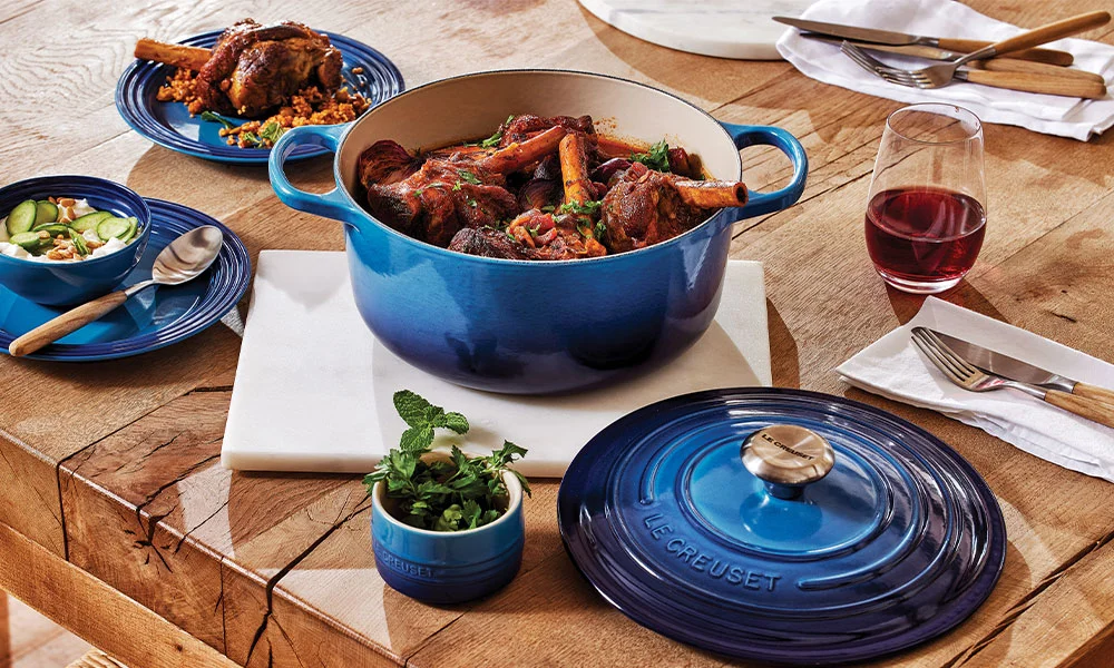 Ein blauer Le Creuset-Bräter mit geschmortem Fleisch auf einem Esstisch, daneben Teller und ein Glas Rotwein.