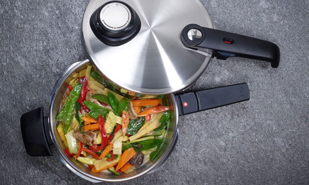 Drucktopf Vitavit von Fissler mit Deckel, gefüllt mit bunten, geschnittenen Gemüse, auf grauem Untergrund.