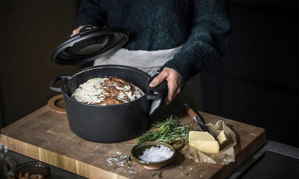 Person öffnet einen gusseisernen Bräter von Fiskars mit frisch gebackenem Brot; daneben frische Kräuter, Meersalz und Butter auf einem Holzbrett.