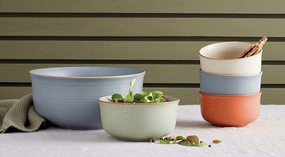 Keramikschüsseln der Serie Nature von Thomas in verschiedenen Farben, gefüllt mit Salat und Brot, auf einem Tisch mit beiger Tischdecke vor grünem Hintergrund.