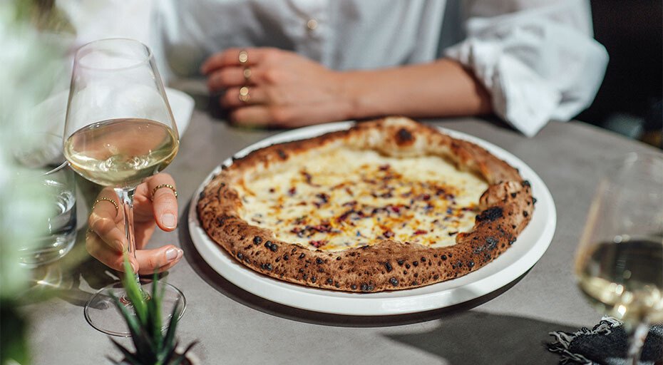 Person mit Weinglas neben einer Pizza auf Tisch, weiße Tischdecke im Hintergrund.