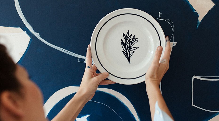Person hält Teller der Serie Solid Color Brasserie von Dibbern mit Pflanzenmotiv vor dunkelblauer, künstlerisch gestalteter Wand.