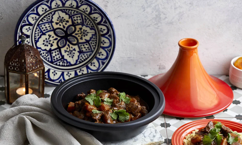 Ein traditionelles marokkanisches Tajine-Gericht in schwarzer Le Creuset Tajine, umgeben von Keramik und dekorativen Elementen.