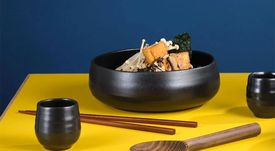 Schwarze Schüsseln mit Tofu, Pilzen und Algen auf gelbem Tisch vor blauem Hintergrund, mit Essstäbchen und Bechern.