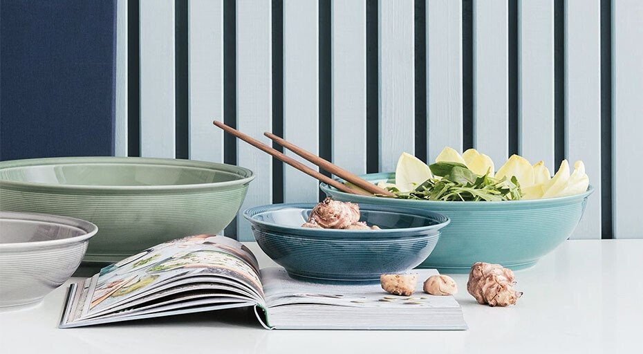 Verschiedene Schüsseln und ein aufgeschlagenes Buch auf weißem Tisch vor geriffelter blauer Wand.
