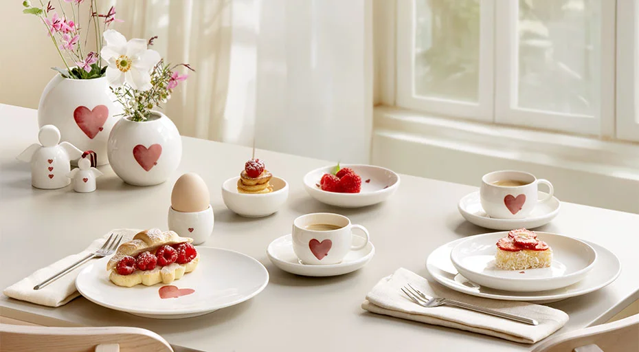 Geschirrset der Serie With Love von Villeroy &amp; Boch mit Herzmotiven, gedeckter Tisch mit Kaffee, Gebäck, Vase mit Blumen, helles Fenster im Hintergrund.