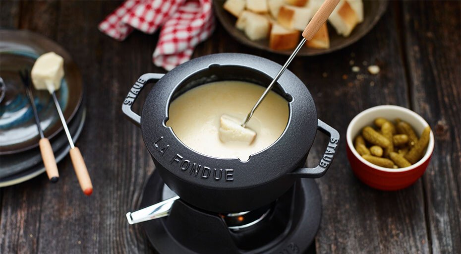 Käsefondue in einem schwarzen Topf mit Brotstücken und einer Schale Gewürzgurken auf einem Holztisch.