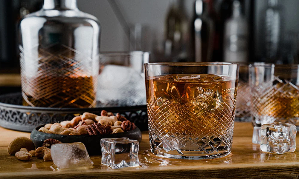 Kristallgläser mit Whisky und Eiswürfeln, umgeben von Nüssen auf einem Tablett.