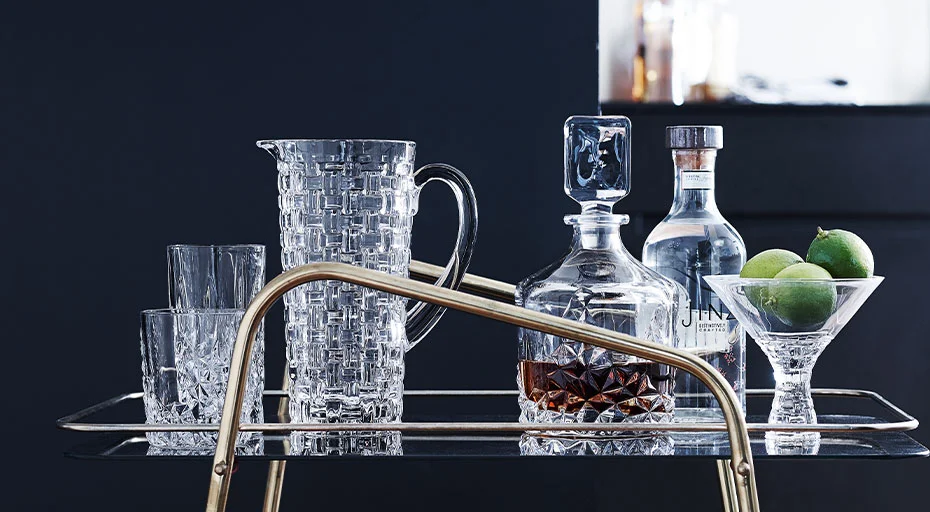 Barwagen mit Kristallkaraffe, Gläsern von Nachtmann, Gin-Flasche und Limetten in einem Schälchen vor dunkelblauem Hintergrund.
