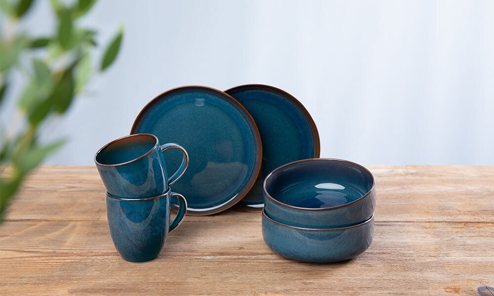 Blaues Geschirr-Set mit Tellern, Schalen und Tassen auf einem Holztisch, unscharfes Pflanzendetail links.