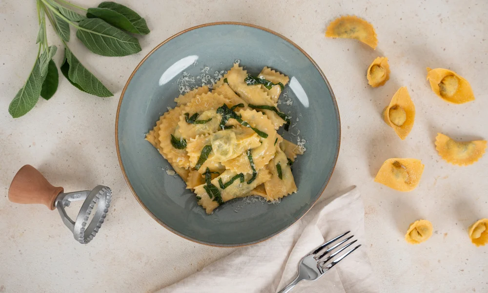 Teller mit Ravioli, daneben liegen einzelne Tortelloni und ein Teigrad auf einem hellen Untergrund.