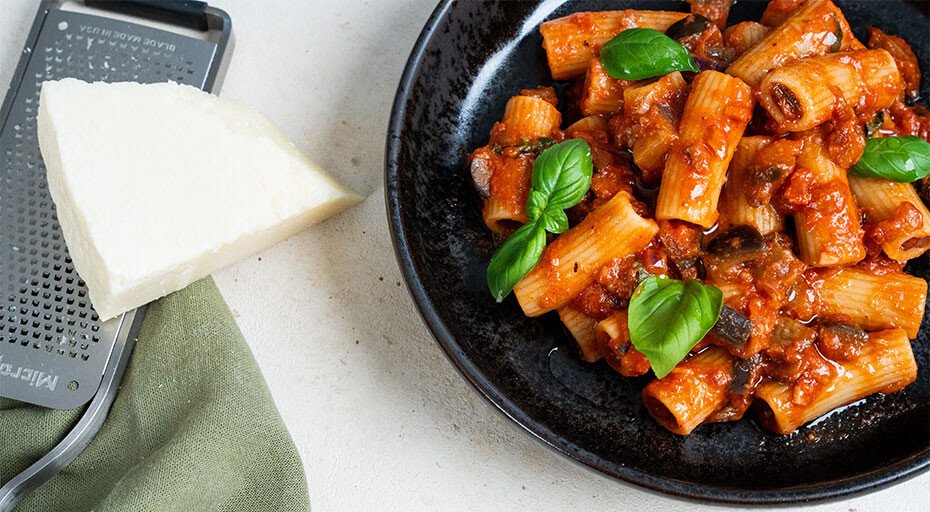 Schwarzer Pasta-Teller mit Rigatoni in Tomatensoße, garniert mit Basilikumblättern; daneben eine Parmesanreibe und Käse.