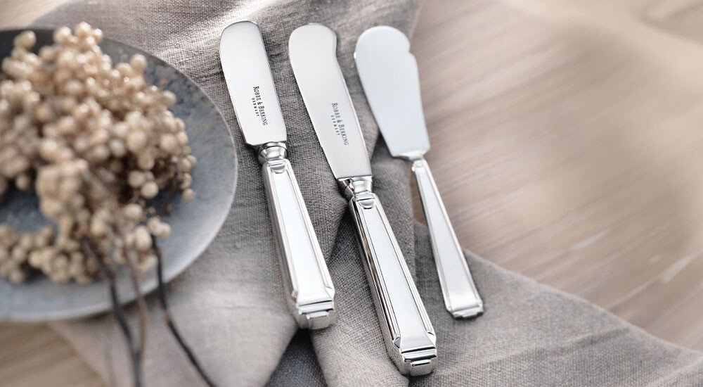 Drei versilberte Buttermesser der Serie Art Deco von Robbe &amp; Berking auf einem hellen Stofftuch neben getrockneten Blumen auf einem Teller.
