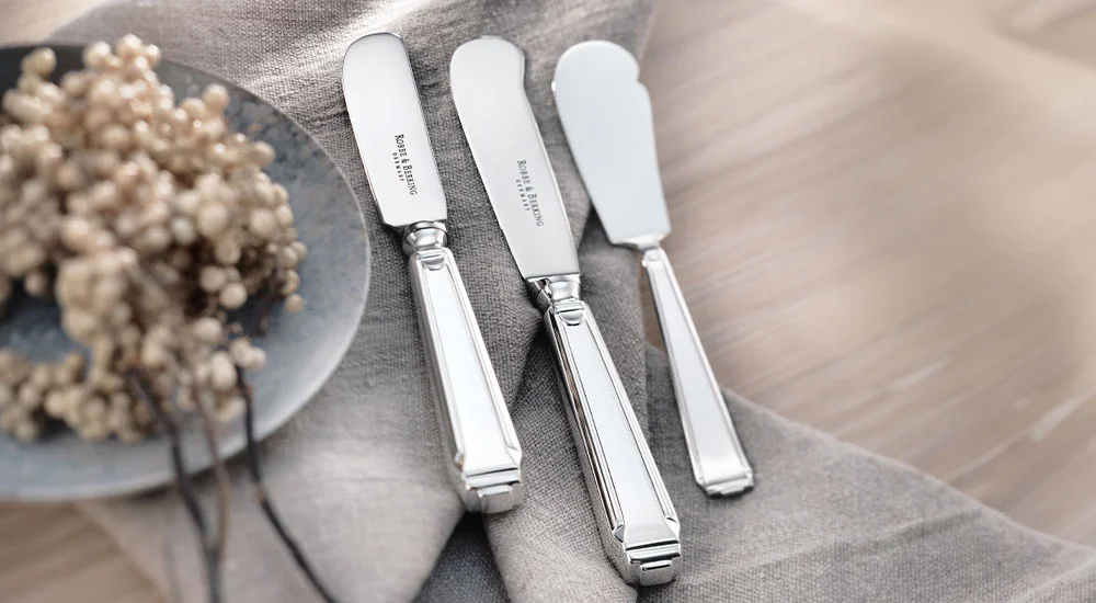 Drei versilberte Buttermesser der Serie Art Deco von Robbe &amp; Berking auf einem hellen Stofftuch neben getrockneten Blumen auf einem Teller.