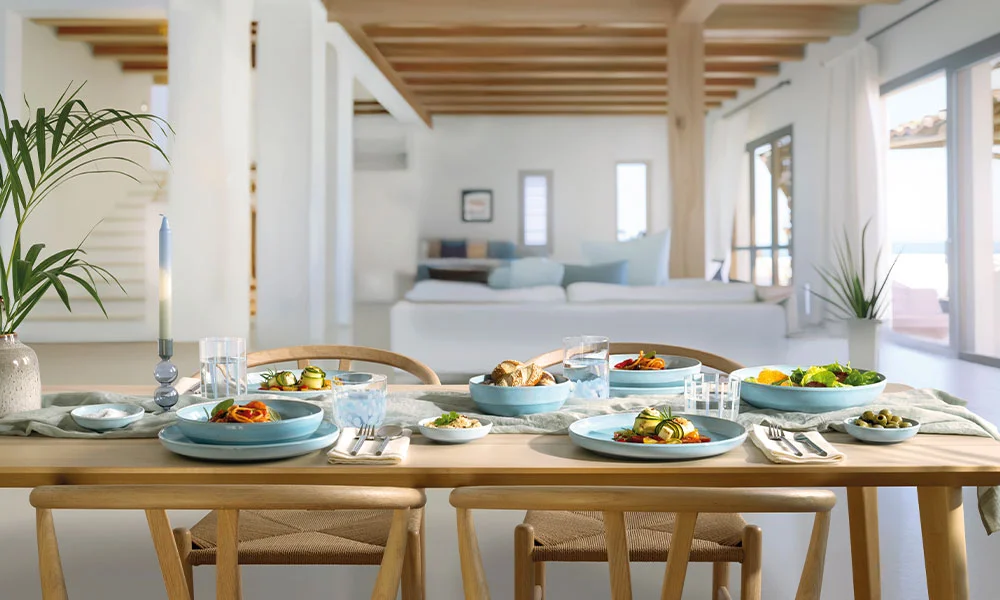 Ein gedeckter Esstisch mit blauem Geschirr der Serie Perlemor Aqua von Villeroy &amp; Boch und Speisen, in einem hellen Raum mit Holzdecke und Pflanzen dekoriert.