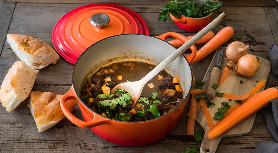Ein orangefarbener Le Creuset Bräter mit Eintopf, Holzlöffel, daneben Brot, Möhren, Zwiebeln und Kräuter auf Holztisch.