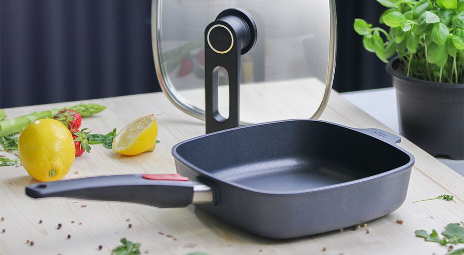 Schwarze quadratische Diamond-Lite-Bratpfanne von Woll mit Deckel auf Holztisch, umgeben von Kräutern, Zitrone und halbierten Tomaten.