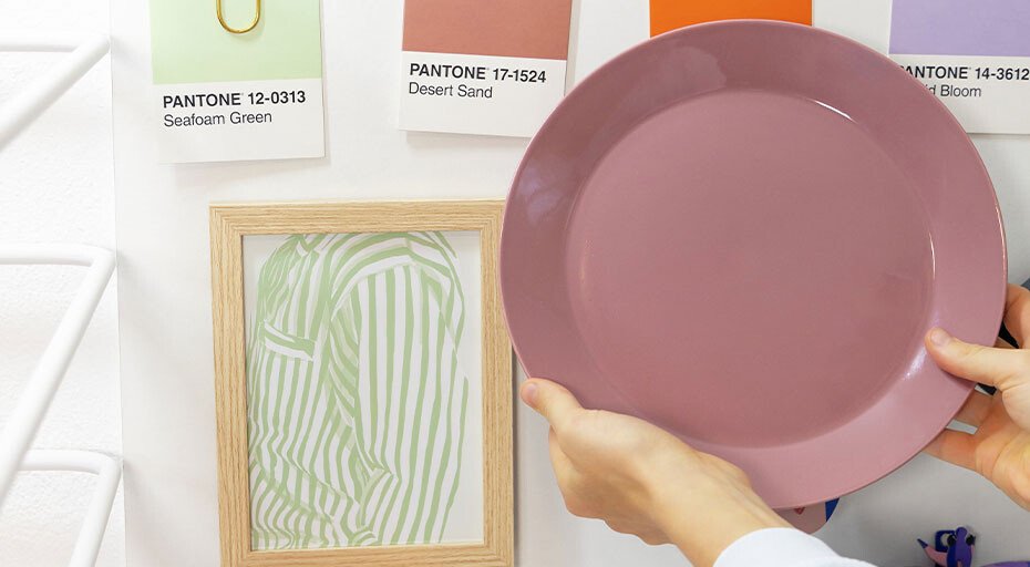 Hand hält einen rosafarbenen Teller vor einer Wand mit Pantone-Farbkarten und gerahmtem Kunstwerk.