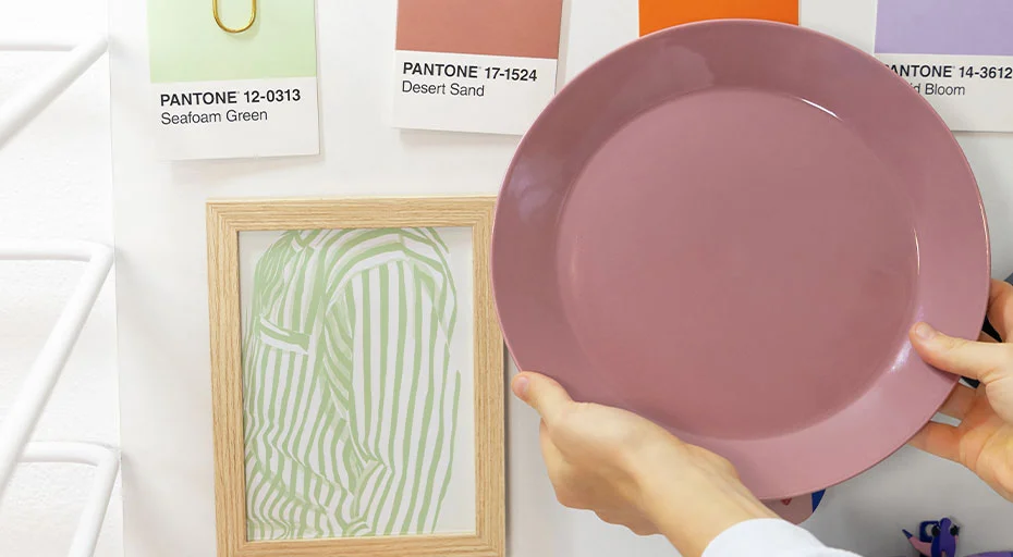 Hand hält einen rosafarbenen Teller vor einer Wand mit Pantone-Farbkarten und gerahmtem Kunstwerk.