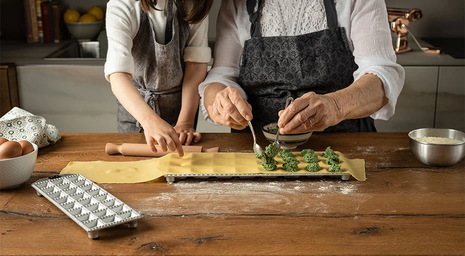 Zwei Personen bereiten Ravioli in einer Küche zu, füllen Teig mit einer Spinatmischung auf einem Holzarbeitsplatz.
