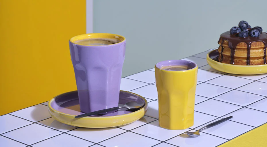Geschirr der Serie Tapero &amp; Ti amo viva von ASA: Lila und gelbe Tassen mit Kaffee, daneben ein Teller mit Pfannkuchen und Blaubeeren auf weiß gekacheltem Tisch.